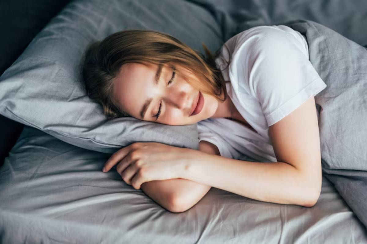 A person with sandy hair lies curled up on a gray bedding, resting their head on a pillow, appearing peaceful and comfortable.