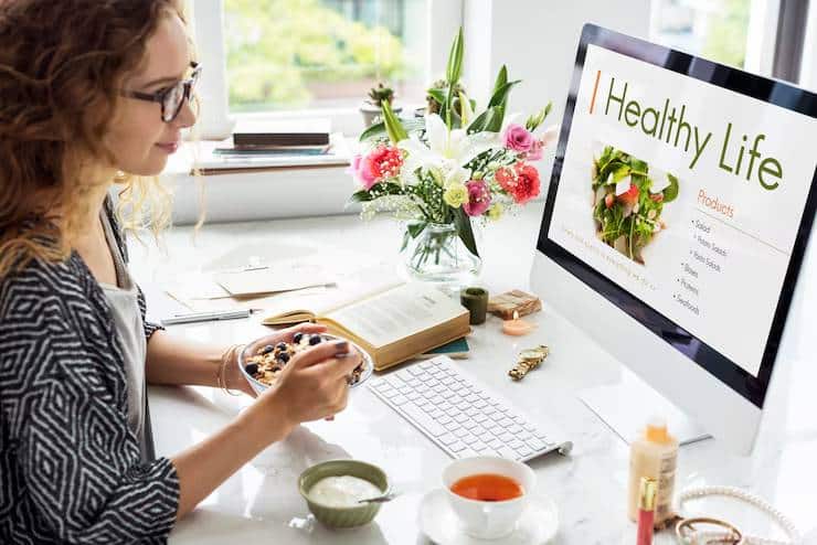 A person enjoys a bowl of healthy food while working at a desk with a computer, a book, and a bouquet of flowers.