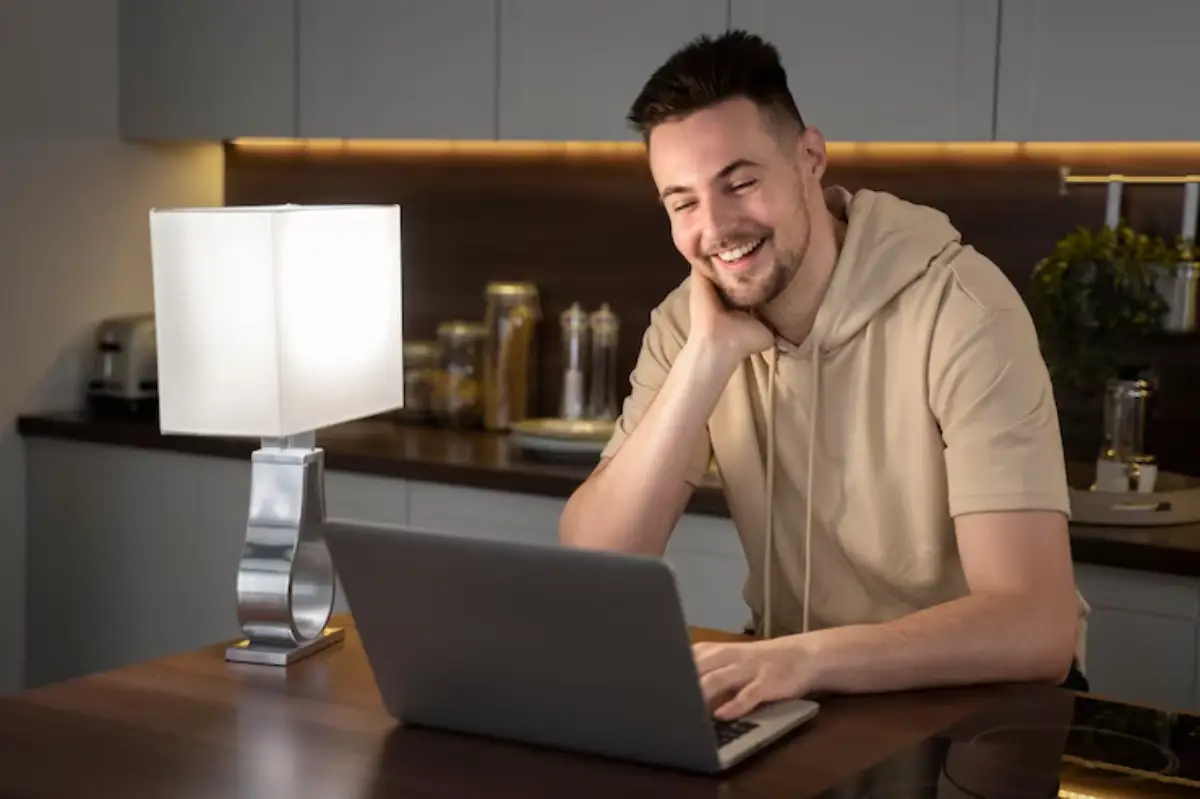  A person in a beige hoodie sits at a wooden table, working on a laptop, with a modern lamp and kitchen items in the background.