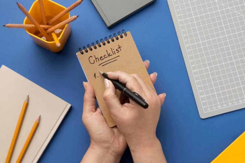 A person's hands holding a notepad labeled Checklist, writing with a pen, surrounded by colored pencils and stationery on a blue surface.