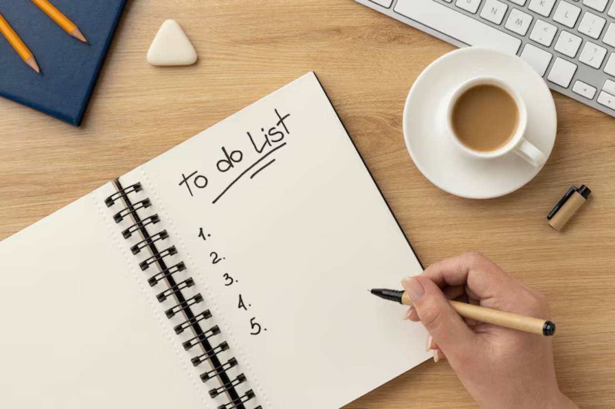  A hand holding a pen writes a to-do list in a spiral notebook, with a cup of coffee and keyboard in the background.