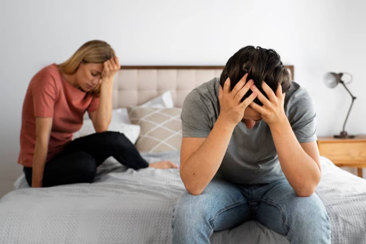 A distressed couple sits on a bed, one with their head in hands, the other looking down, indicating tension or conflict. 
