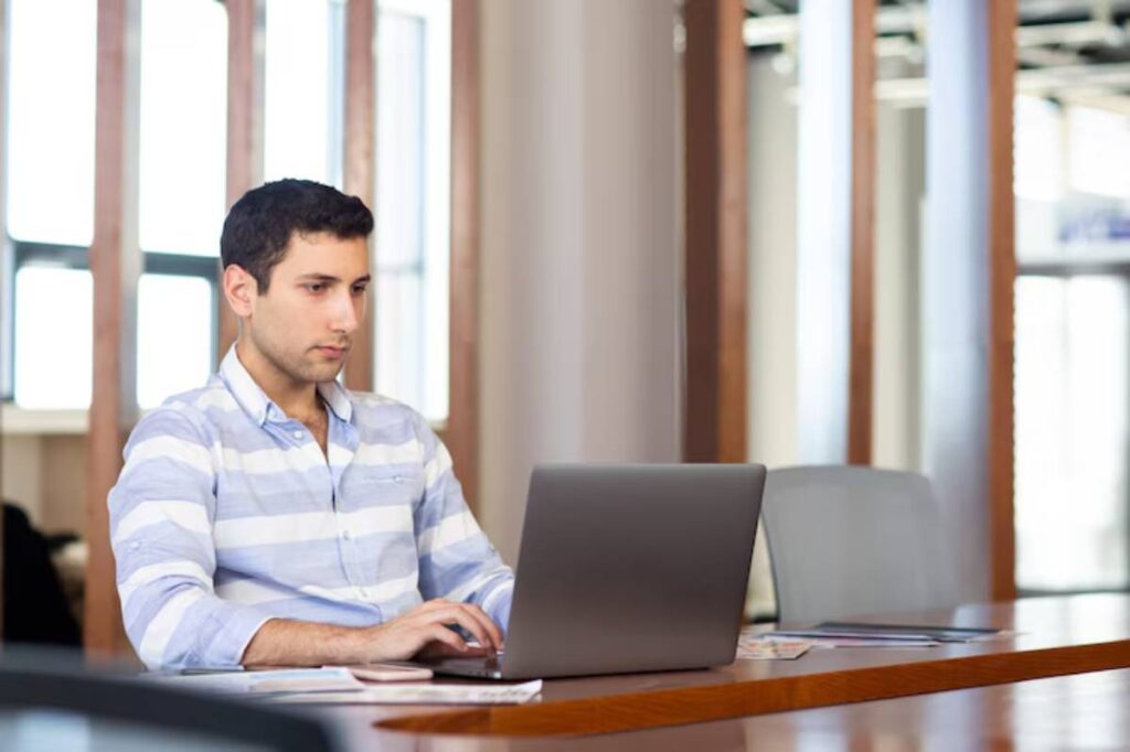A person in a striped shirt works on a laptop at a desk, surrounded by large windows and modern office furniture.