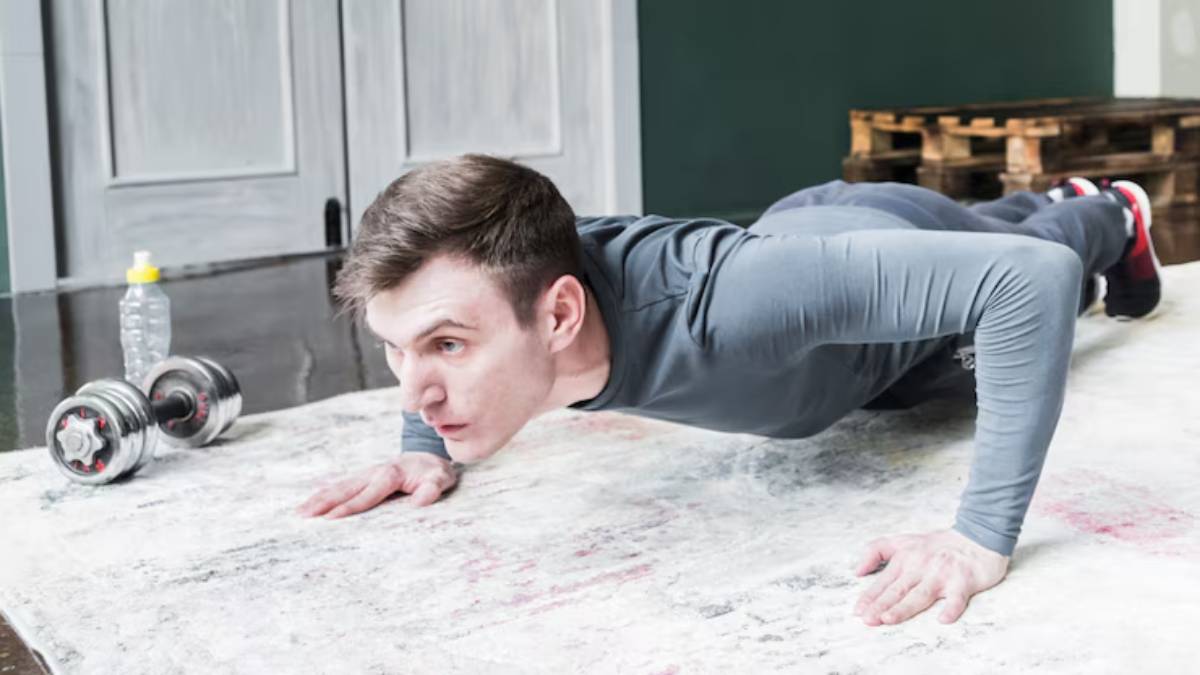 A person performs push-ups on a rug, with dumbbells and a water bottle nearby in a modern indoor setting.