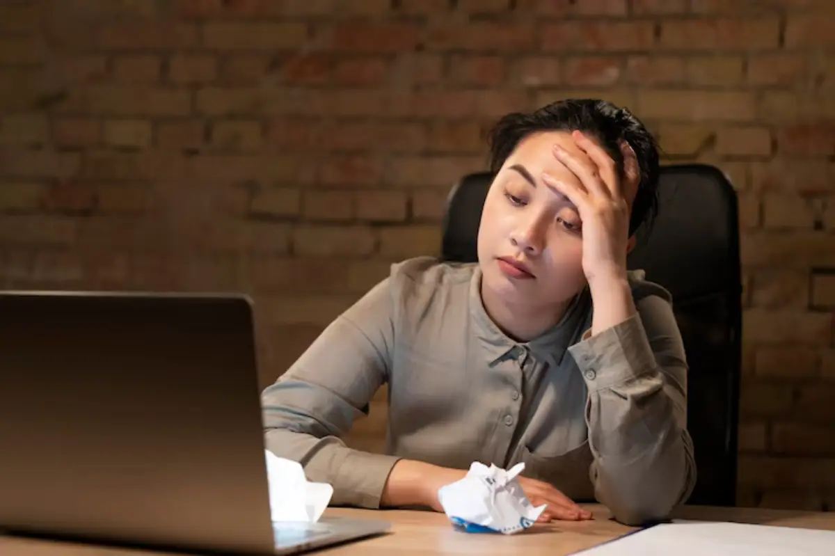  A person in a gray shirt rests their chin on their hand, surrounded by crumpled paper on a desk with a laptop against a brick wall.