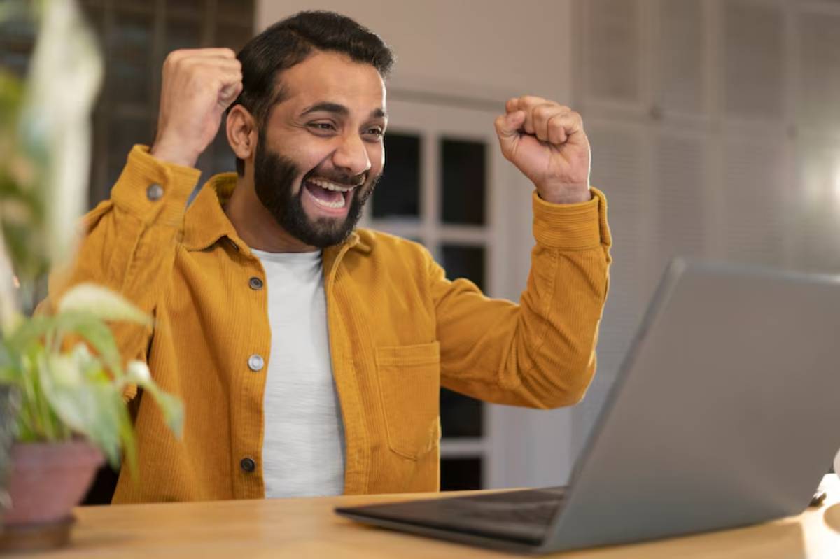 A person in a yellow jacket celebrates joyfully in front of a laptop, with arms raised in excitement, in a cozy indoor setting.