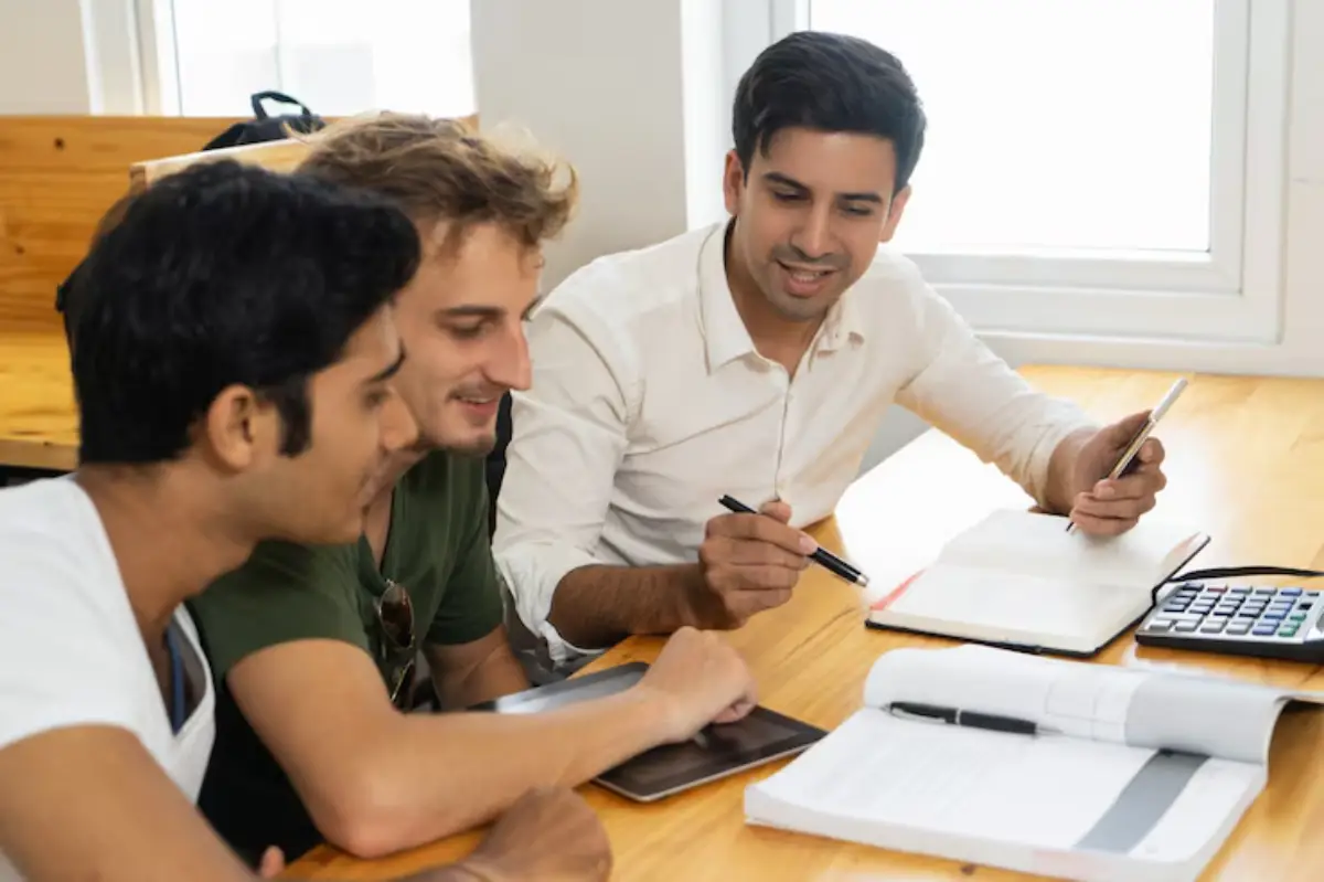  Three students engage in a collaborative study session at a wooden table, surrounded by notebooks, a tablet, and a calculator.