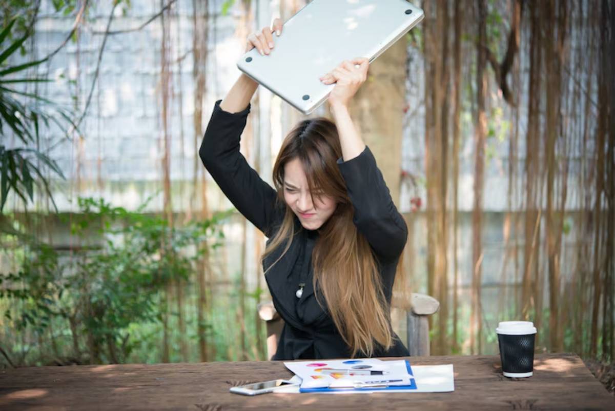 A person raises a laptop above their head in frustration, surrounded by plants, a coffee cup, and scattered papers on a wooden table.