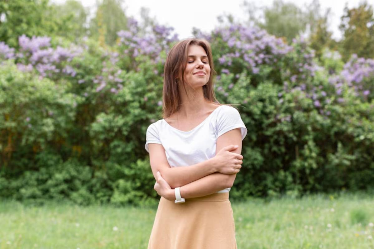 A woman stands in a lush green field, arms crossed, wearing a white shirt and a beige skirt, with colorful flowers in the background.