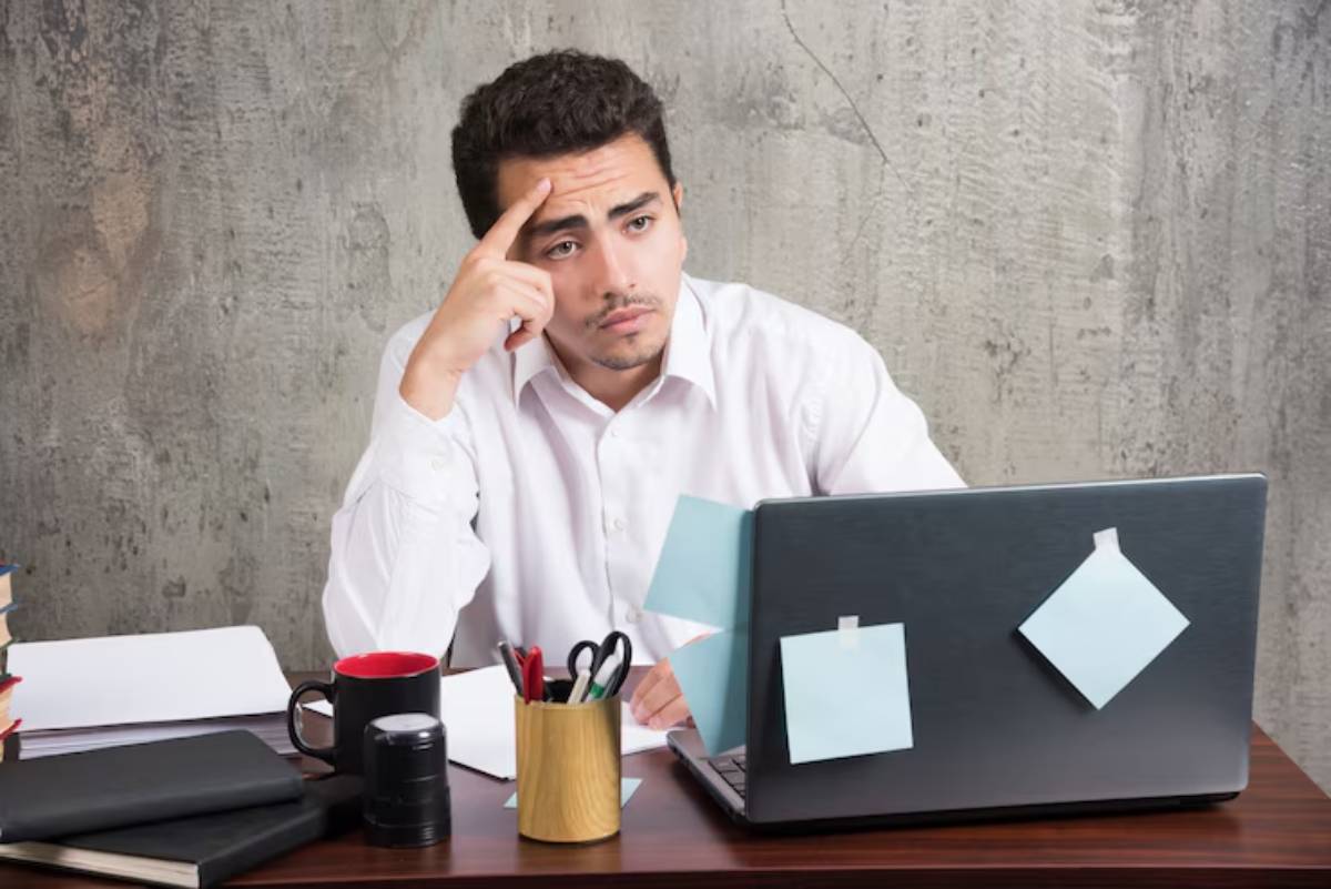 A man in a white shirt sits at a desk with a laptop, looking stressed, surrounded by notebooks, a mug, and office supplies.