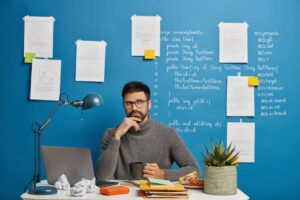 A person in a gray sweater sits at a desk cluttered with papers and a plant, against a blue wall filled with notes and code.