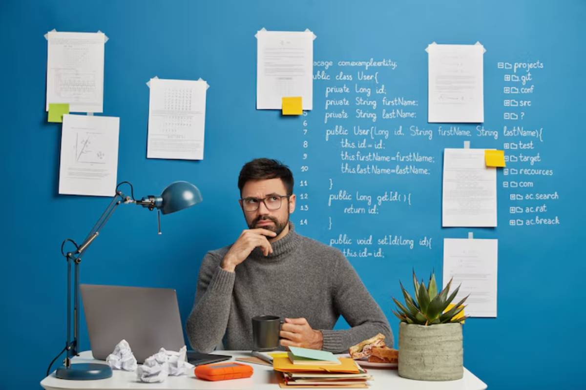 A person in a gray sweater sits at a desk cluttered with papers and a plant, against a blue wall filled with notes and code.