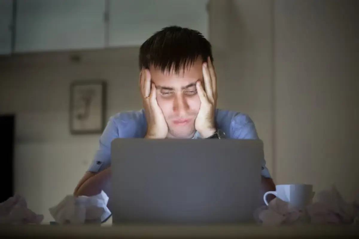  A person sits in front of a laptop with hands on their head in a dimly lit room, surrounded by crumpled tissues and a cup.