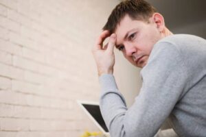 A person in a gray sweater resting his head on his hand, seated in front of a laptop against a light brick wall.
