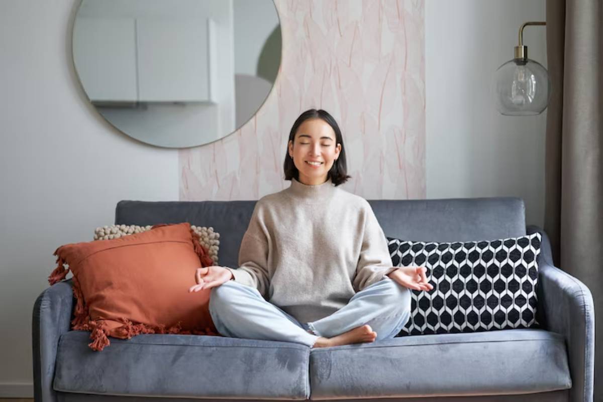 A person sits cross-legged on a gray couch surrounded by decorative pillows, meditating in a cozy living room setting.