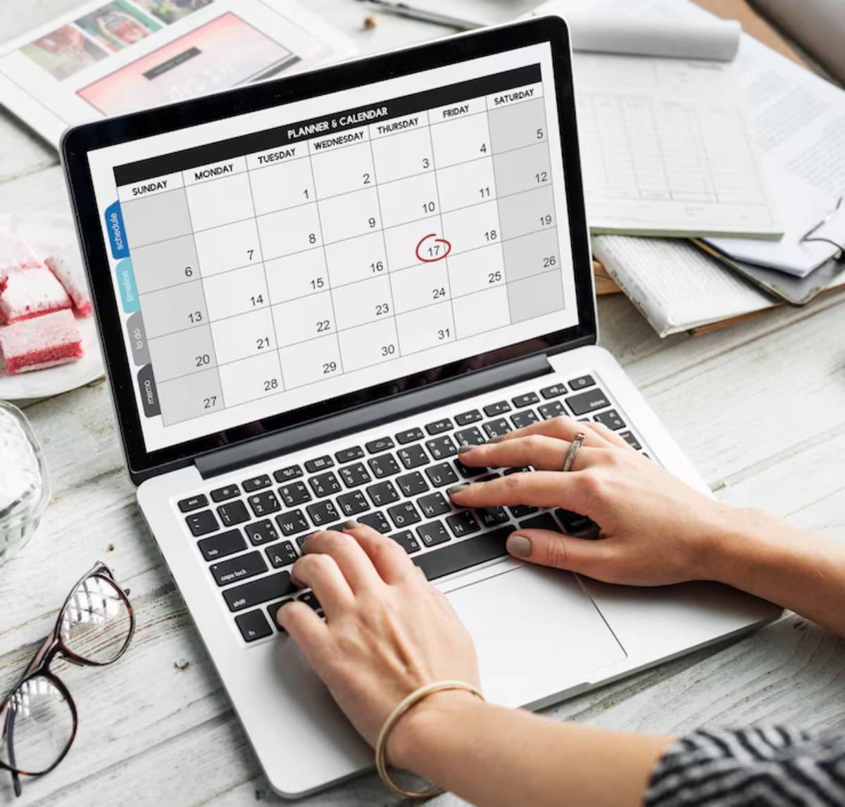  A person views a digital calendar on a laptop, highlighting the 17th, with notebooks and snacks on a wooden table.