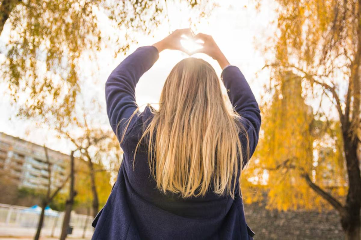  A woman with long hair creates a heart shape with her hands against a bright sky, surrounded by autumn trees and a park backdrop.