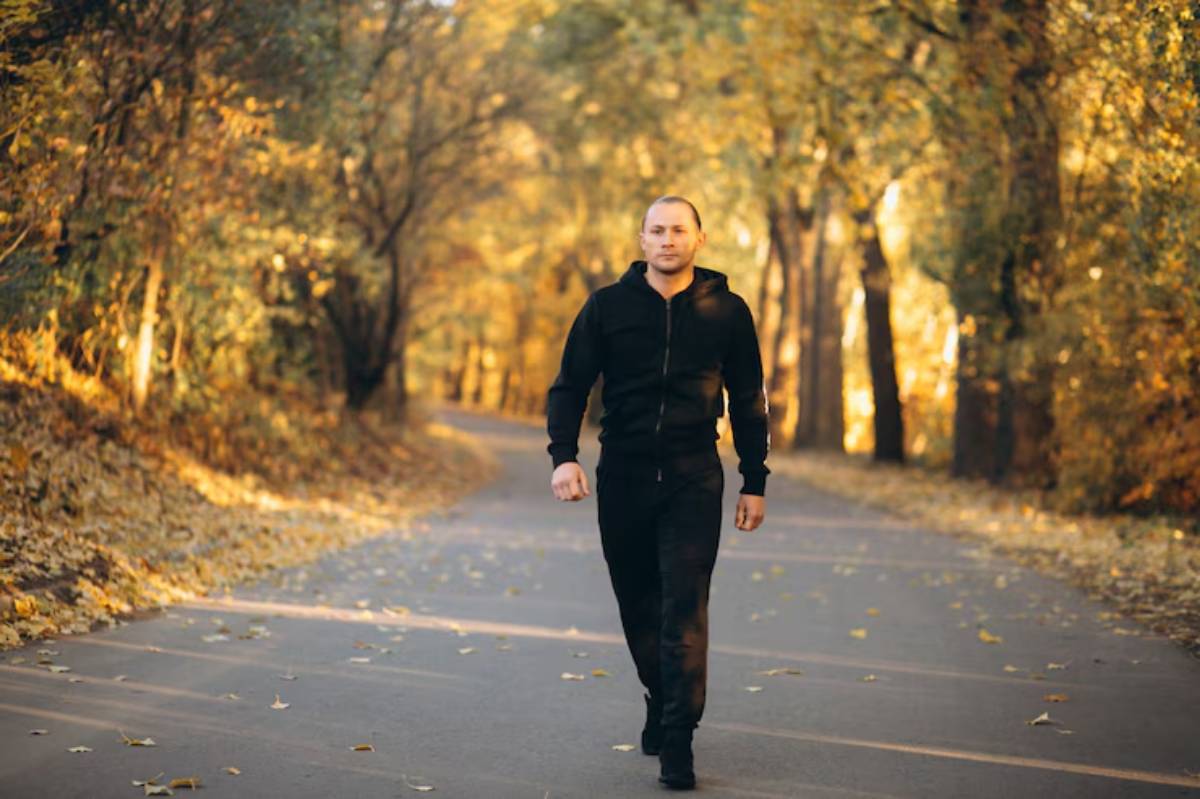 A person in a black outfit walks down a picturesque tree-lined path covered in autumn leaves, bathed in warm, golden light.
