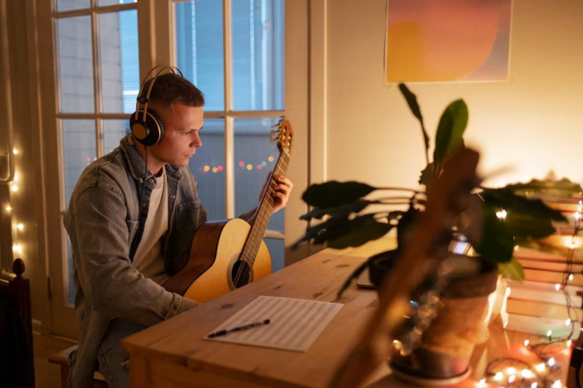 A musician plays an acoustic guitar while wearing headphones, surrounded by soft lighting and a cozy, creative workspace.