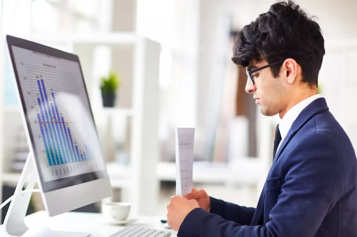  A man in a suit is reviewing a report while looking at a computer screen displaying various charts in a bright office setting.