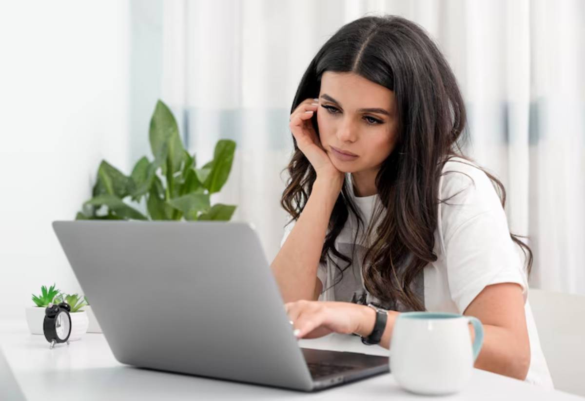 A person sits at a desk, focused on a laptop, with a potted plant and a coffee cup nearby, in a bright indoor setting.