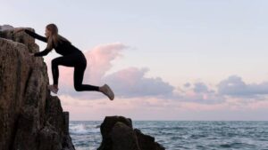 A person in black clothing climbs a rocky shoreline with the ocean and pastel sky in the background during twilight.