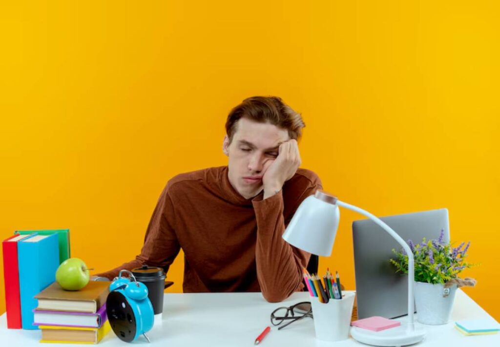 A person rests their head on their hand at a desk with books, a lamp, an apple, and a laptop against a bright yellow background.