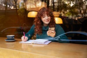 A person with curly hair sits at a cafe table, writing in a notebook and using a smartphone, with a coffee cup beside them.