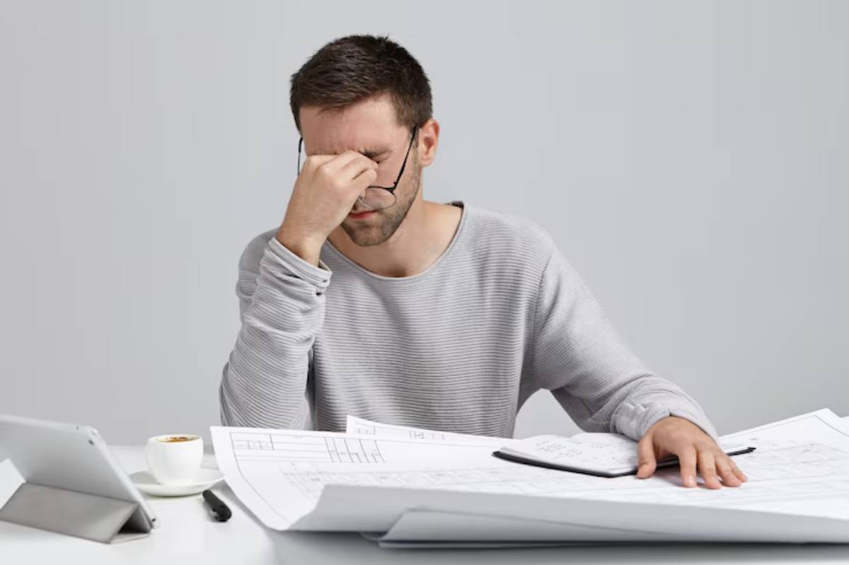  A person in a gray sweater rests their chin on their hand, surrounded by blueprints, a notebook, and a cup of coffee on a table.