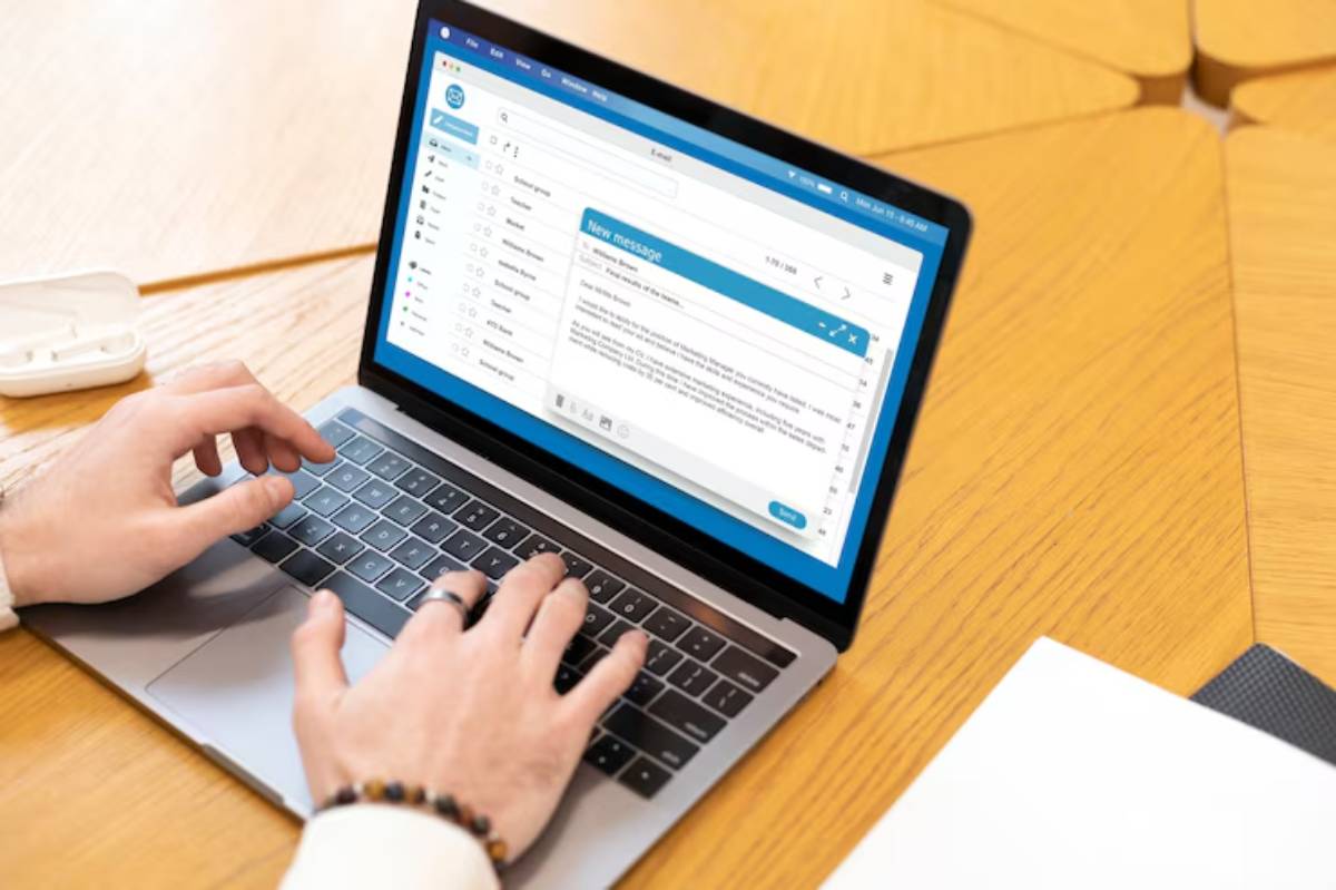 A person typing an email on a laptop, with an open email interface displayed, sitting on a wooden table. 