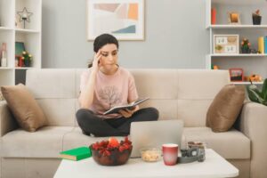 A person sits cross-legged on a couch, reading a book with snacks and a laptop on a coffee table in a cozy living room.