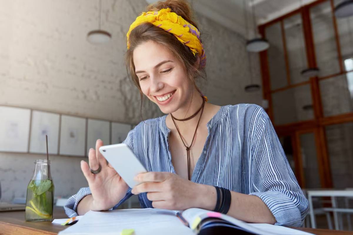A person sits at a table with a notebook, using a smartphone, wearing a blue striped shirt and a yellow headband in a bright, airy space. 